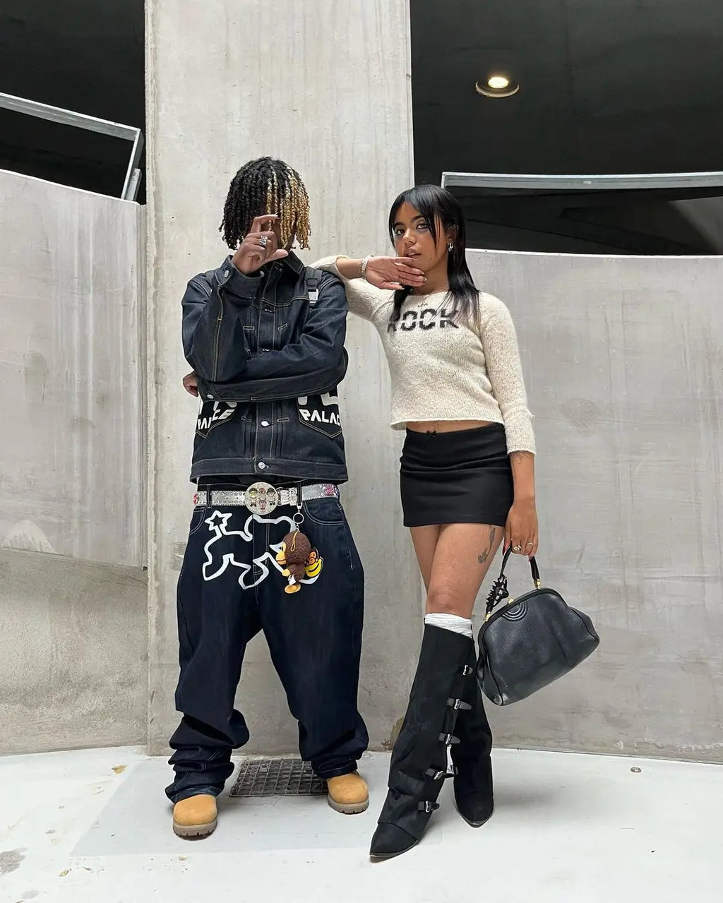 Two people standing against a concrete wall, one wearing a dark outfit with a belt and the other in a light sweater and black skirt.